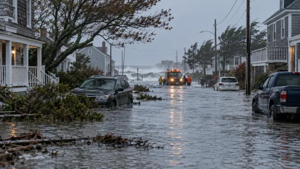 Uma rua inundada num bairro costeiro após uma tempestade de inverno, com veículos submersos e equipas de emergência a trabalhar sob a luz cinzenta da manhã.