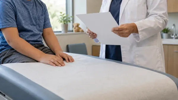 An anonymous child sits in a generic pediatric clinic while a clinician holds a blank sheet during an assessment.