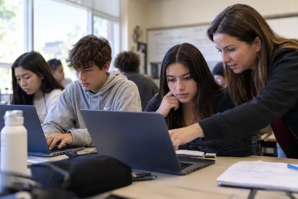 A teacher works with students using laptops and notebooks in a classroom, illustrating supervised technology use in education.