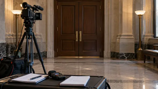 A television camera, two notebooks and an audio recorder positioned outside closed wooden doors in a civic building.