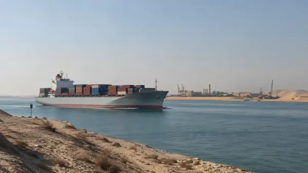 Un buque portacontenedores sin distintivos atraviesa el Canal de Suez a la luz del día, pasando junto a la infraestructura portuaria situada al borde del desierto