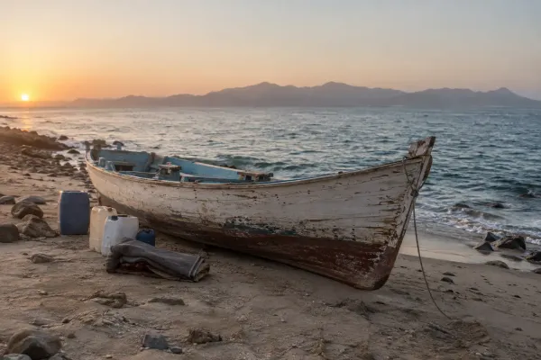An unmarked wooden passenger boat beside water containers and blankets on an arid shoreline at dawn.