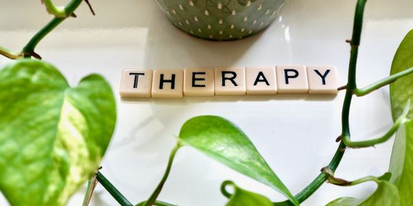 Scrabble letters laid out on a white table spelling the word therapy with some green plants.