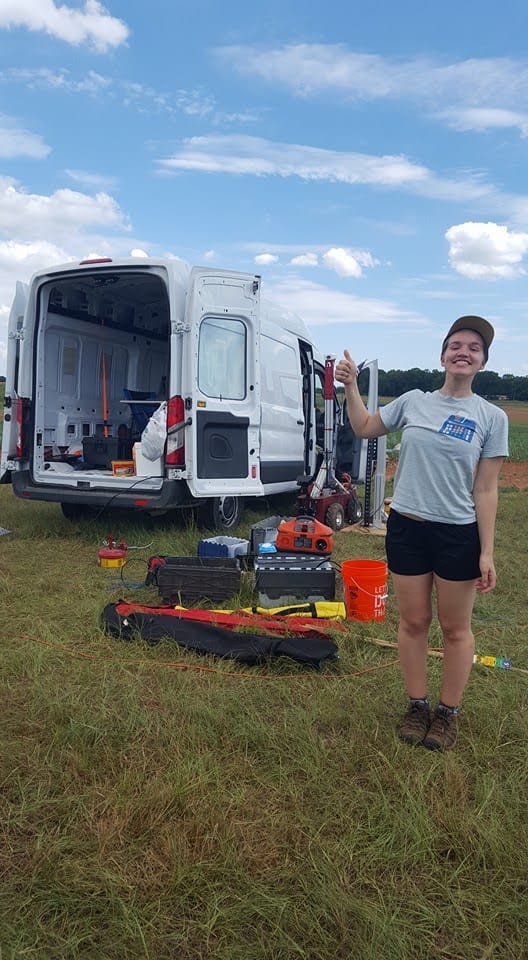 Maggie gives a thumbs up in a muddy field, surrounded by chaotic electronics and supplies. 