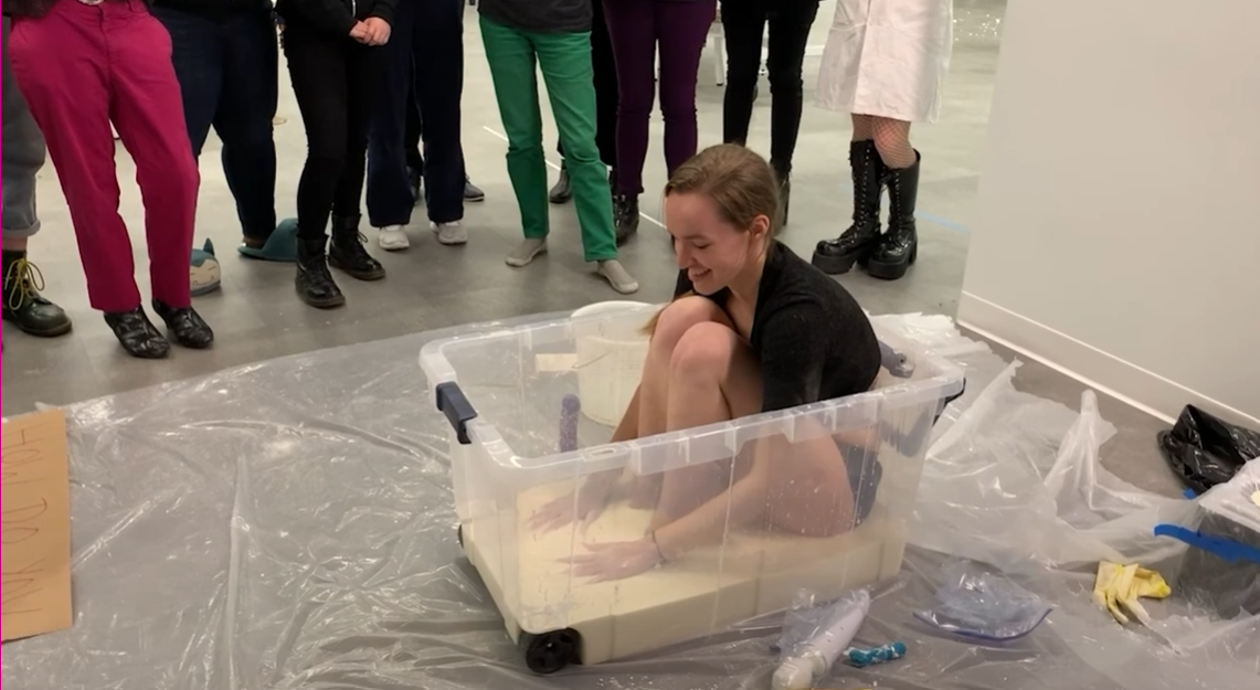 Maggie sits in a plastic vat of ooblek, smiling in underwear. An audience looks on. 