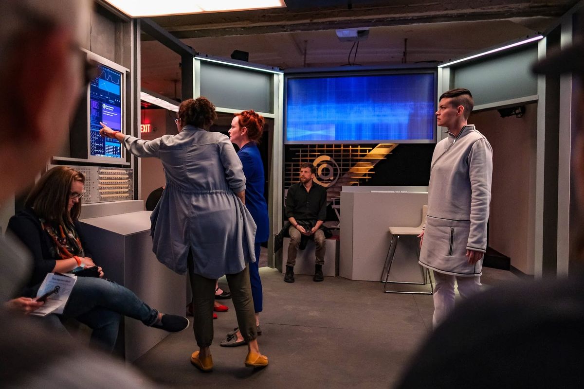 3 actors look at a screen in Project Amelia. One android, one engineer, and one journalist. Audience members watch on.