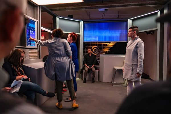 3 actors look at a screen in Project Amelia. One android, one engineer, and one journalist. Audience members watch on.