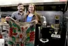 Aethereal Coffee owners Troy McGlashan and Molly Kincaid hold up a hand-painted bag from one of their coffee growers in Nicaragua. Behind them is their coffee roaster in the commercial kitche