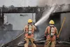 A Central Mat-Su Fire Department firefighter sprays water on a house at the corner of Sullivan Avenue and Julie Marie Circle Thursday afternoon while a member of the Butte Fire Department wai