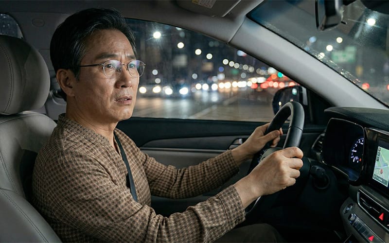 An elderly patient wearing glasses sitting in the driver's seat at night with a worried expression.