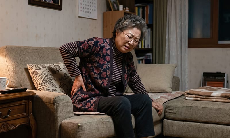 An elderly patient sitting on a couch at night, holding their lower back in discomfort
