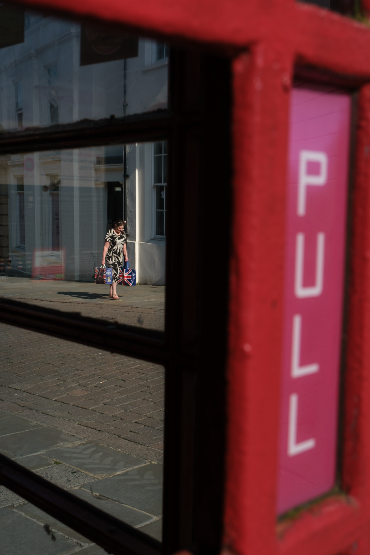 A person is seen through the glass panes of a red structure, likely a phone booth, with a PULL sign visible in close-up. The individual outside is carrying bags adorned with the Union Jack design and is walking on a cobblestone