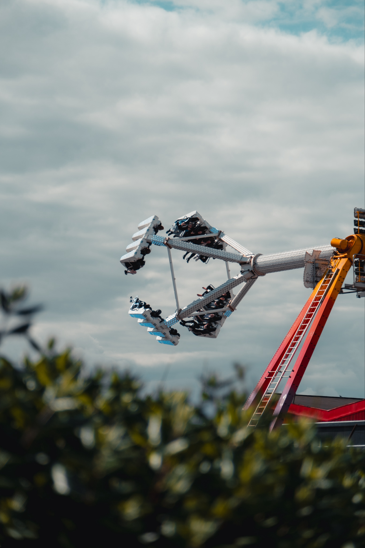 A thrilling amusement park ride with riders in mid-air is set against a cloudy sky, partially obscured by green foliage.