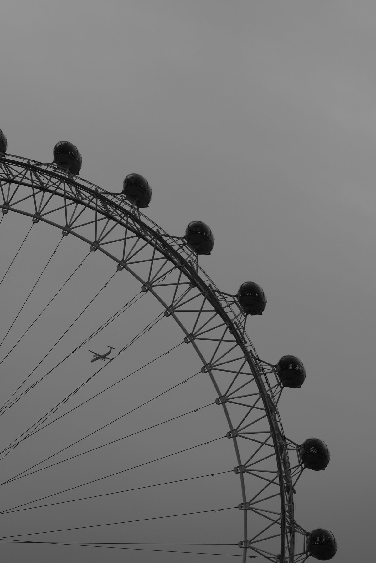 A black and white photograph of a large Ferris wheel, partially visible, with a small airplane in the background sky.