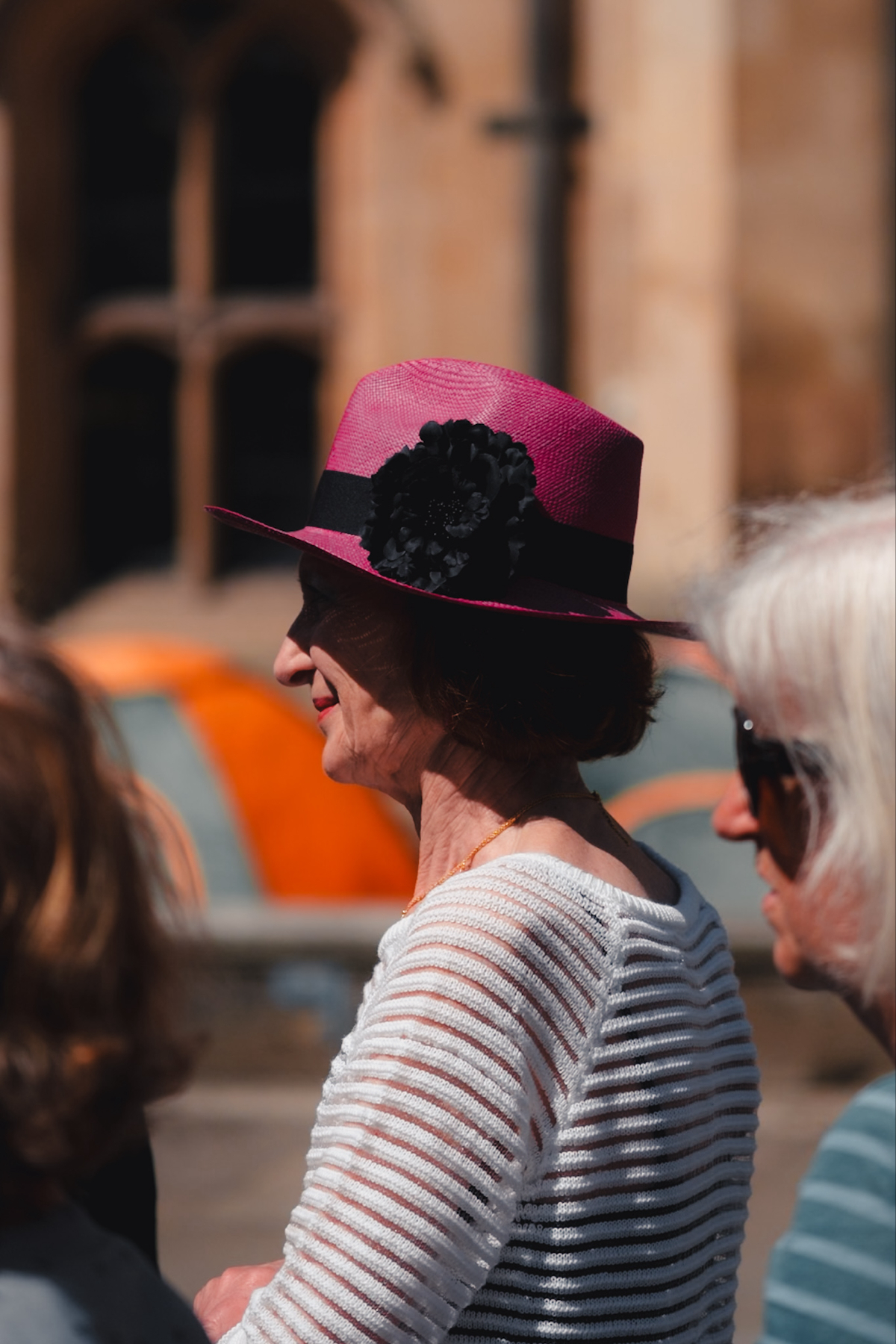 A person wearing a bright pink hat with a black flower and black band, along with a white knit top, standing outdoors beside two others. The background features a blurred architectural structure and some vibrant orange elements.