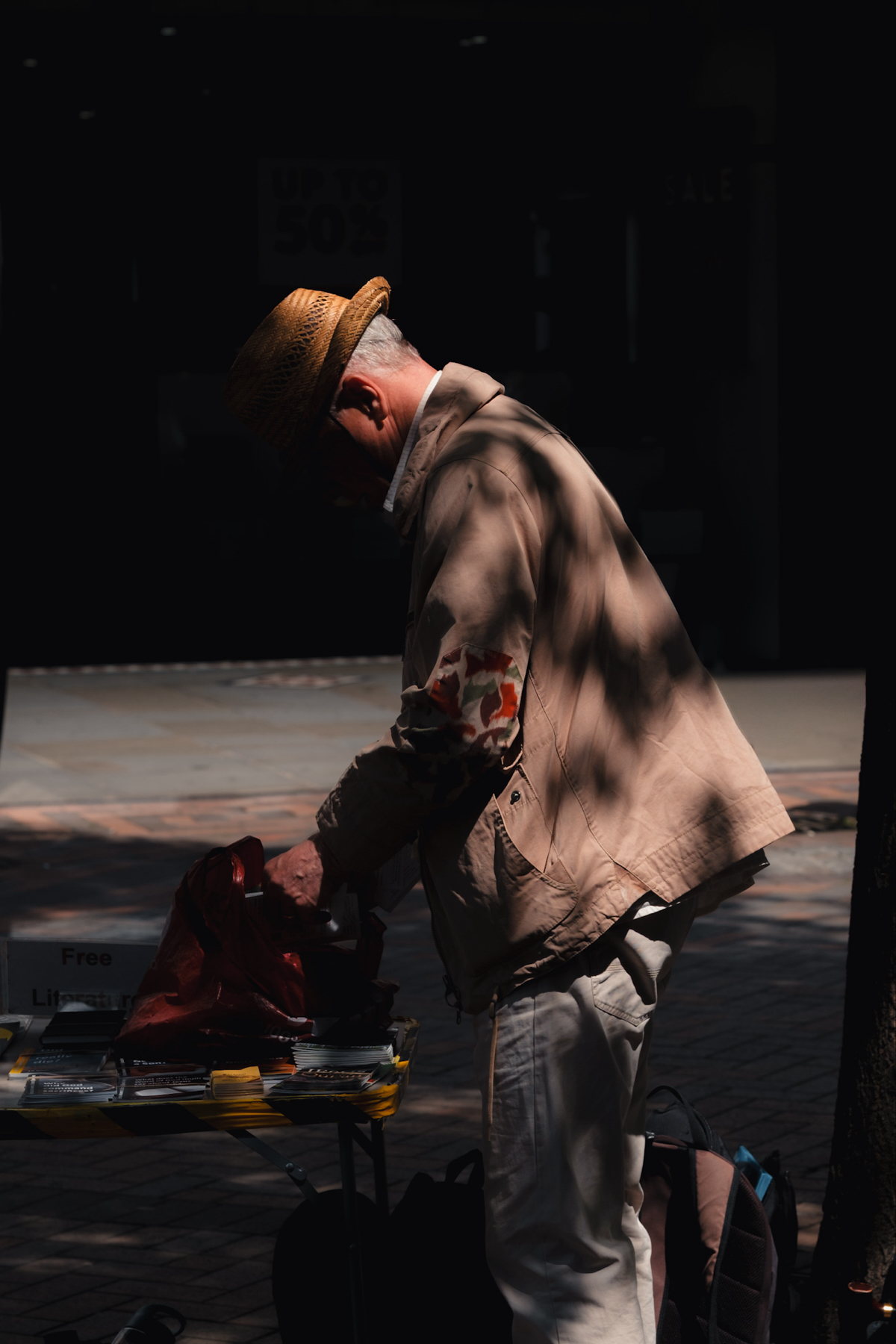 An elderly person wearing a hat and a light jacket, standing by a table with pamphlets labeled "Free Literature." The individual is holding a red bag and appears to be picking up an item from the table. The setting is outdoors in a shaded area