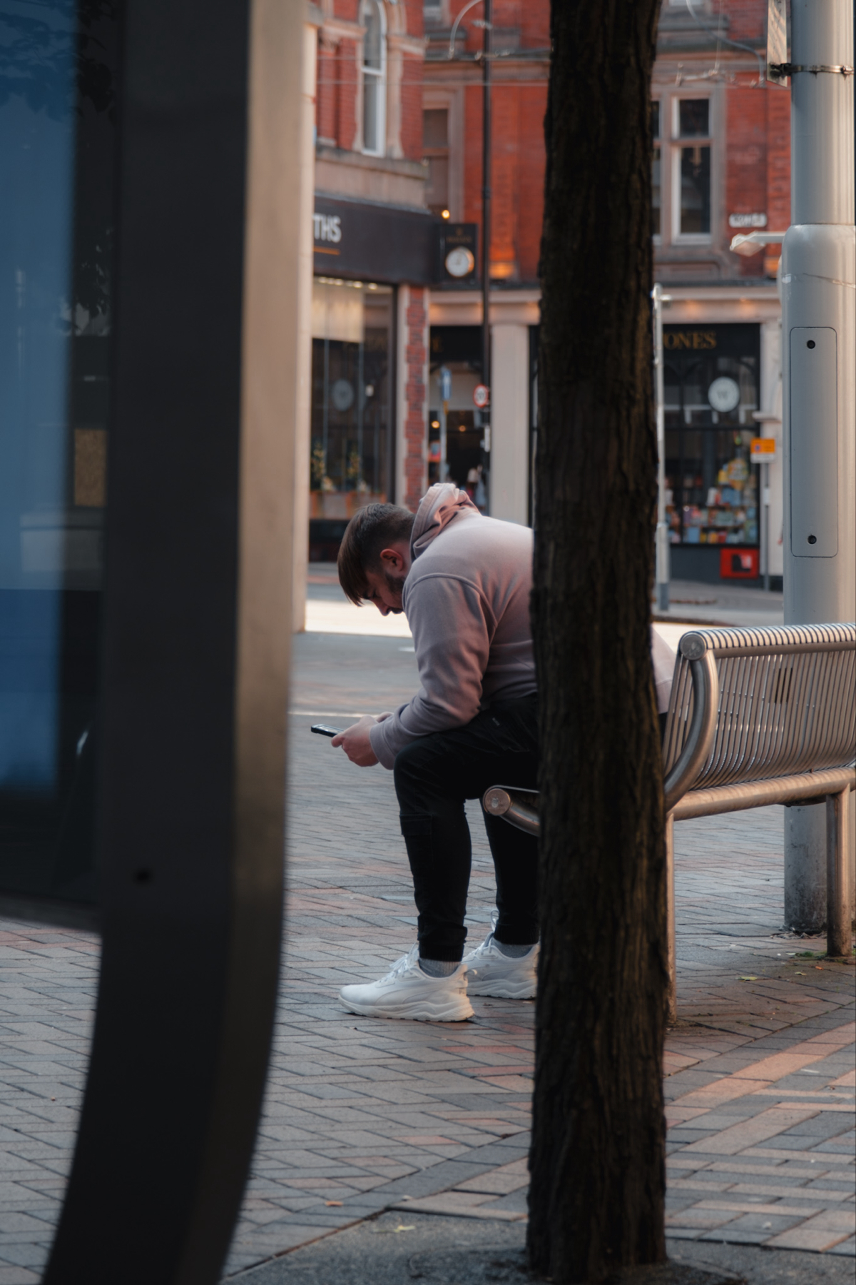A person wearing a hoodie and sneakers sits on a bench, looking at their phone. The setting appears to be an urban area with brick buildings and storefronts in the background. A tree partially obscures the view of the person.