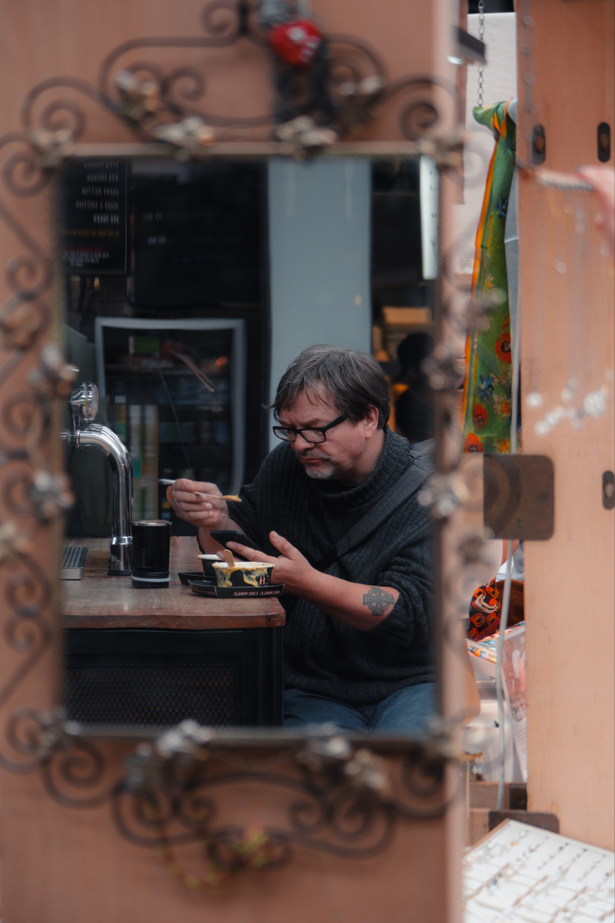 A man with glasses and a tattoo on his left arm is eating at a wooden table, captured through a decorative mirror. He is using a fork and looking down at his food, with a drink and what appears to be a food container in front of