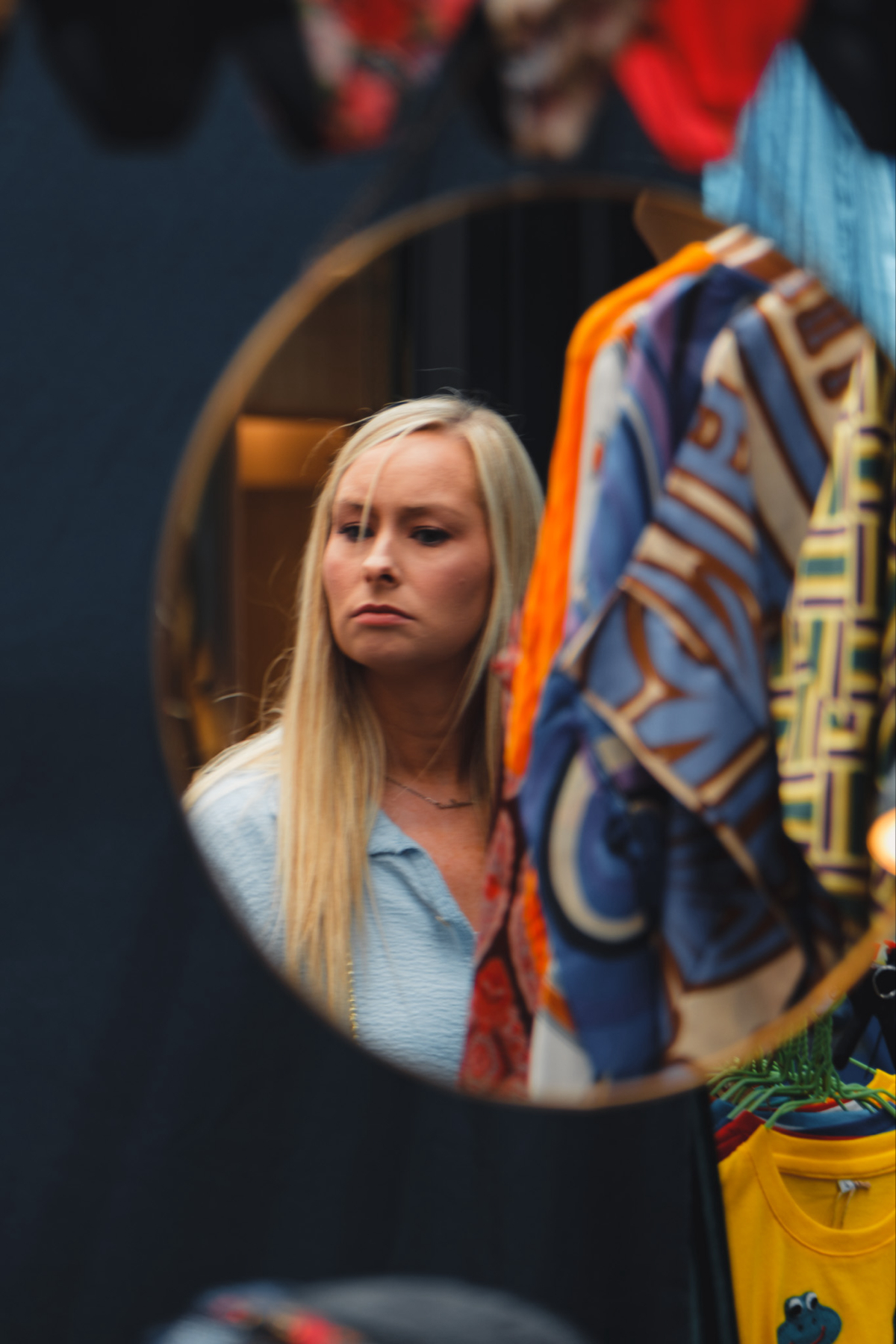 A blonde woman is looking at her reflection in a round mirror, seemingly inspecting clothes off-frame. Various colorful clothing items hang in the foreground, obstructing part of the scene. The background is blurred but appears to be indoors.