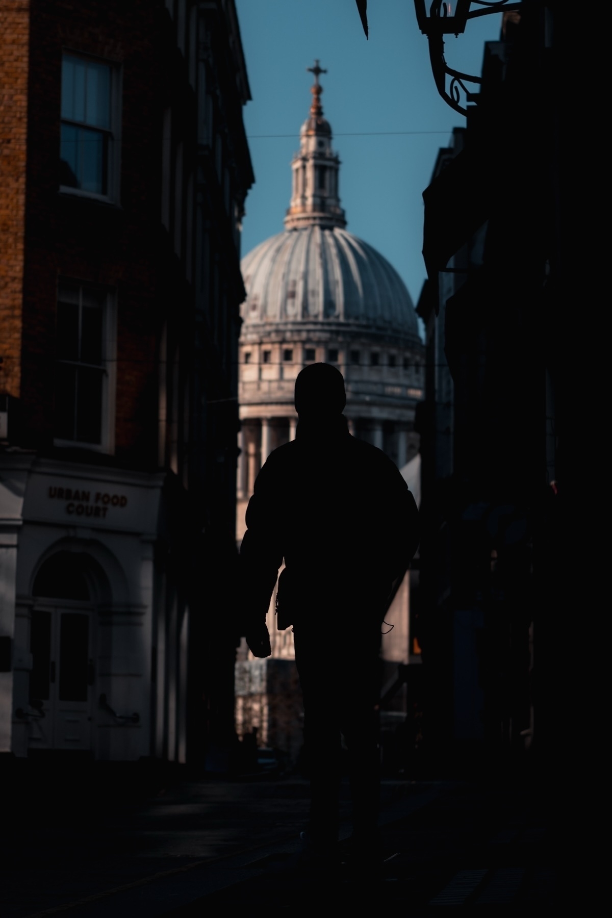 Silhouette of a person walking through an alley with a view of a large domed building in the background, possibly St. Paul's Cathedral. The surrounding buildings create a frame around the dome, emphasizing its architectural prominence.