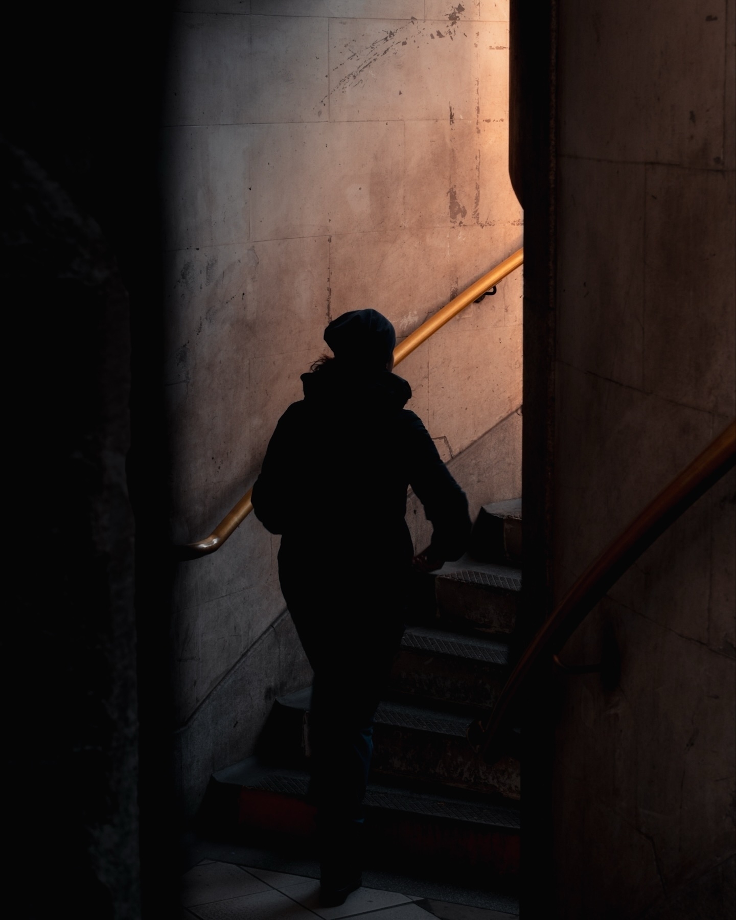 Silhouette of a person ascending a dimly lit staircase with a handrail, set against a textured wall.