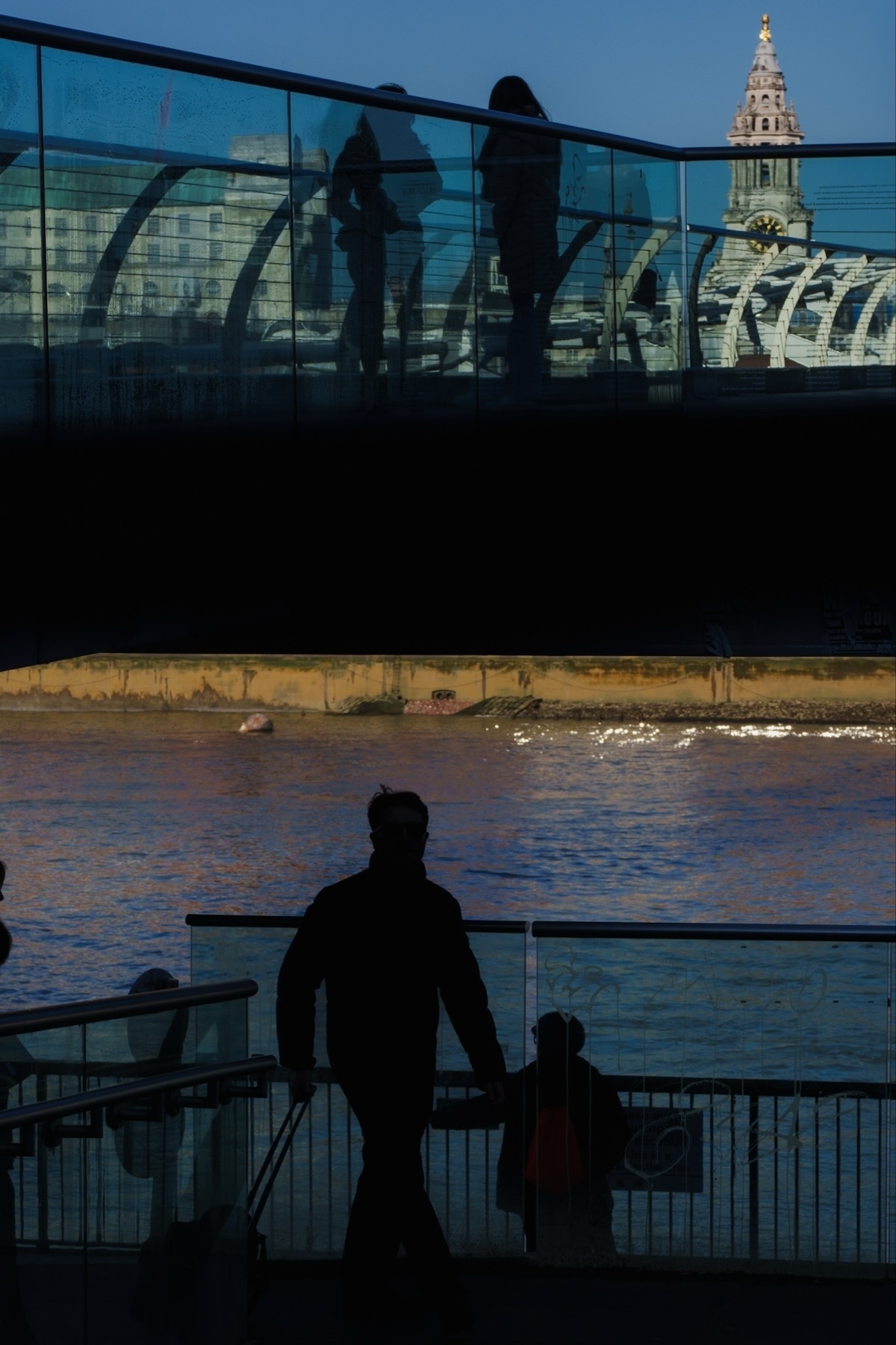 A silhouette of a person walking up a staircase, beneath a glass bridge with reflections of people and buildings visible. In the background, a river and a historic clock tower are illuminated by sunlight.