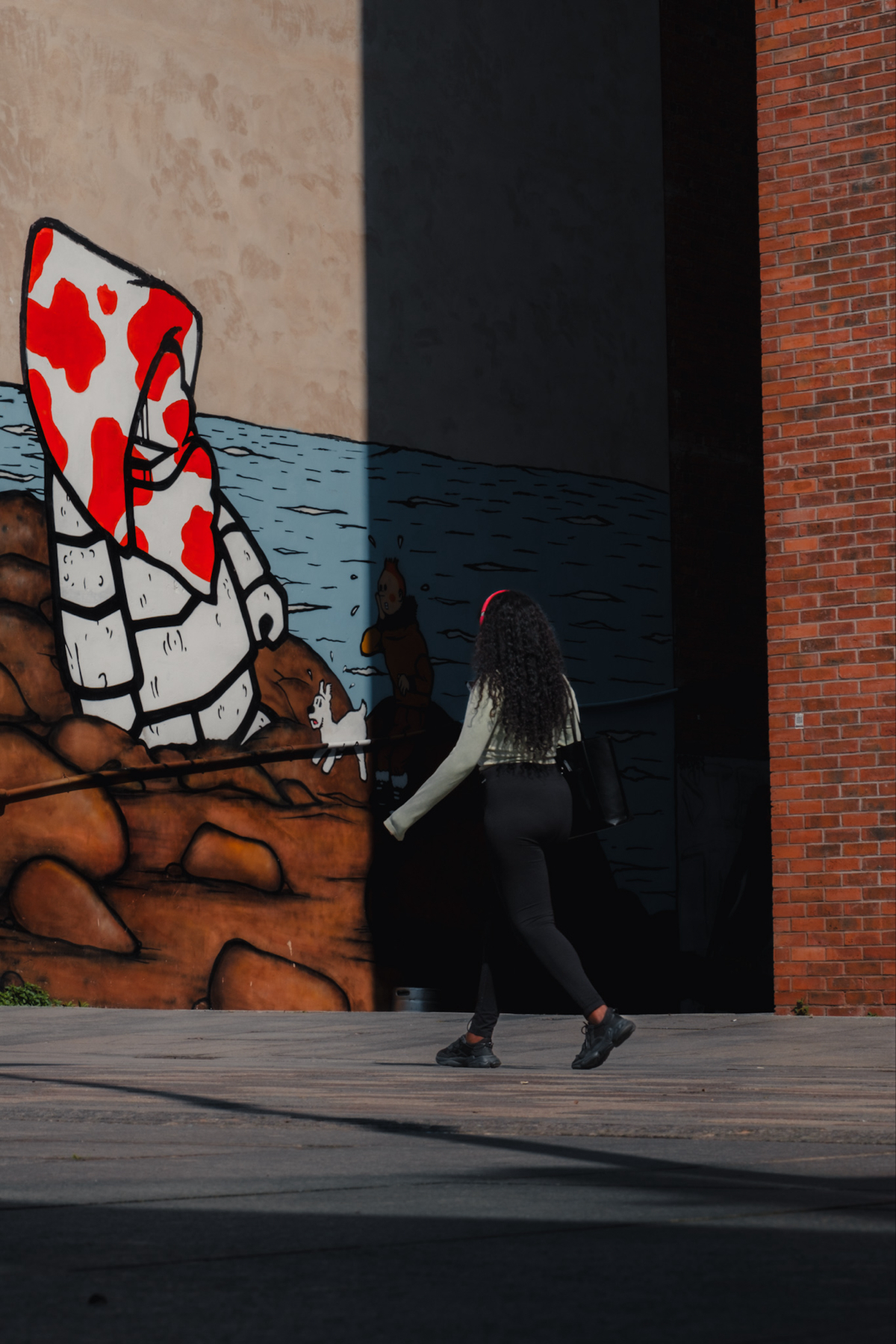 A person walks past a mural on a brick wall. The mural depicts a cartoonish scene with rocks, water, and large figures. The individual in the foreground has dark, curly hair and is wearing a light-colored top, black pants, and black