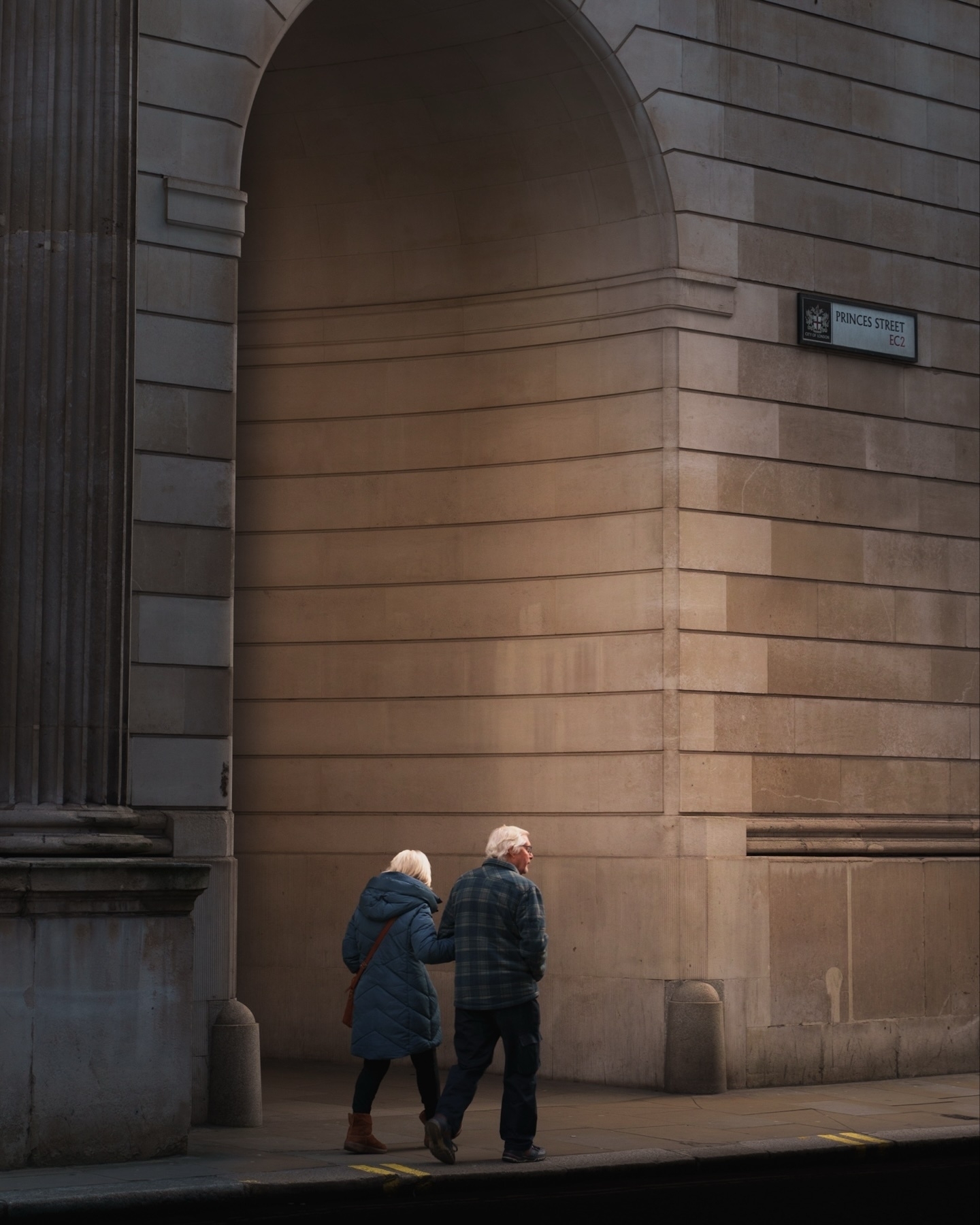 An elderly couple walks hand in hand along a sidewalk near a large stone building with an archway. The building has a sign reading "Princes Street EC2." The scene is calm and the lighting is soft.