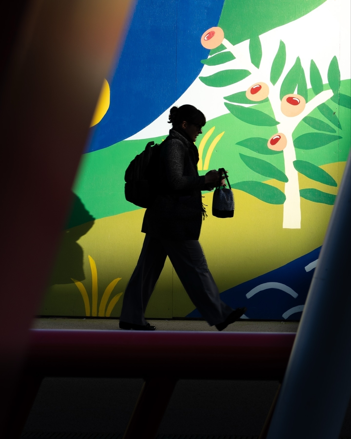 A silhouette of a person walking against a colorful mural featuring abstract designs with leaves, berries, and vibrant shapes. The person carries a backpack and a small bag, with the scene framed by blurred foreground elements.