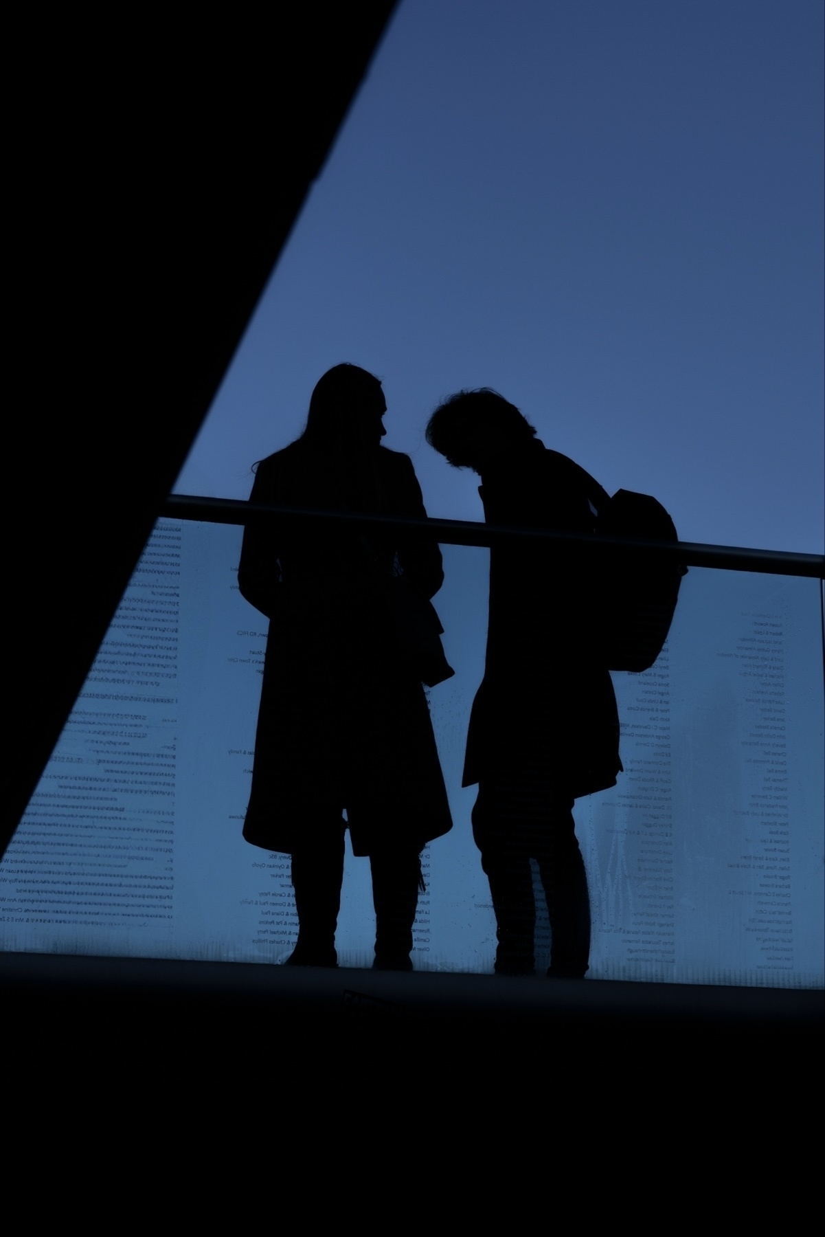 Silhouette of two people standing on a balcony against a blue sky. One person has a backpack. They appear to be engaged in a conversation.