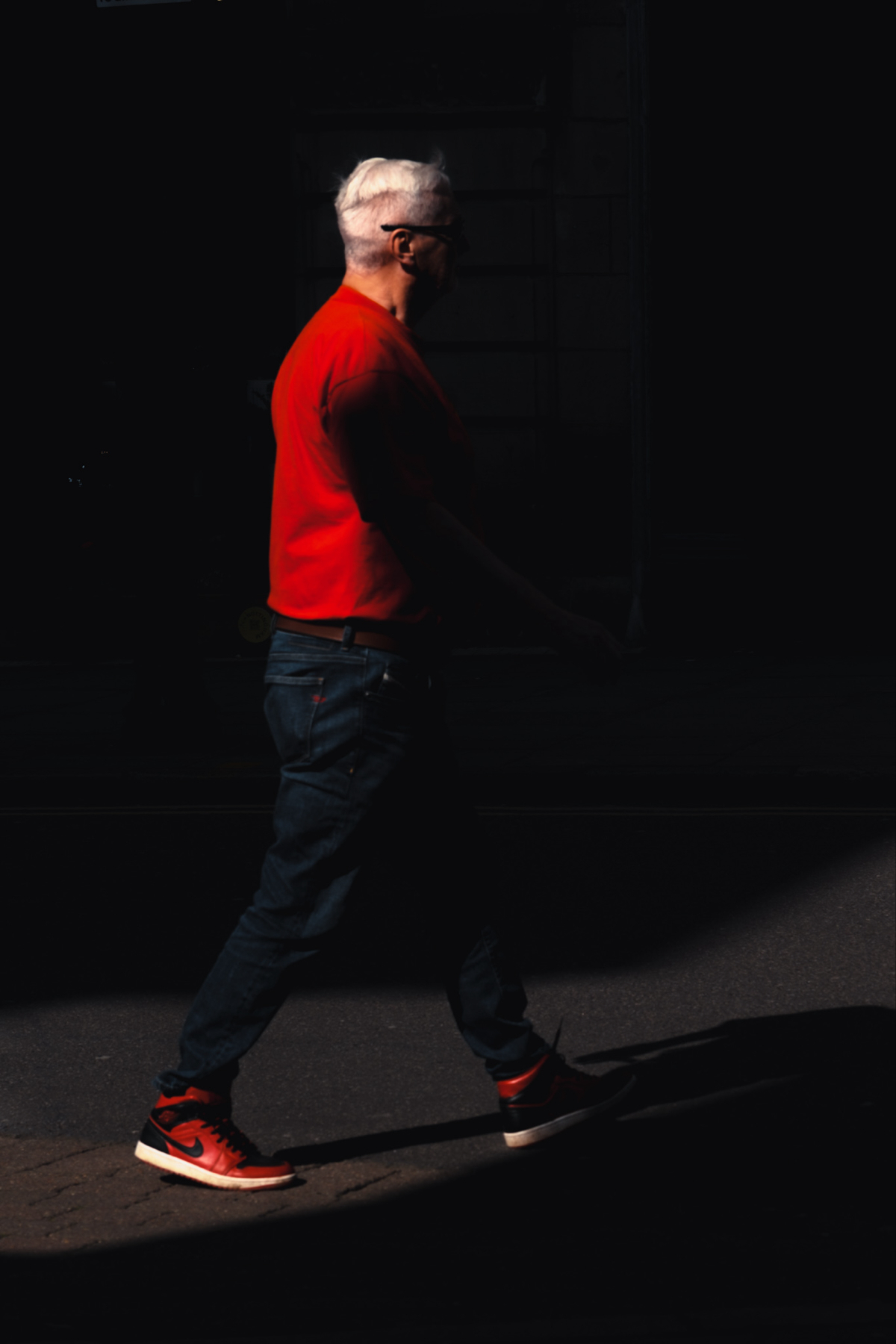An older man with white hair is walking outdoors. He is wearing a red shirt, jeans, and red sneakers. The lighting casts a sharp contrast, highlighting him against a dark background.