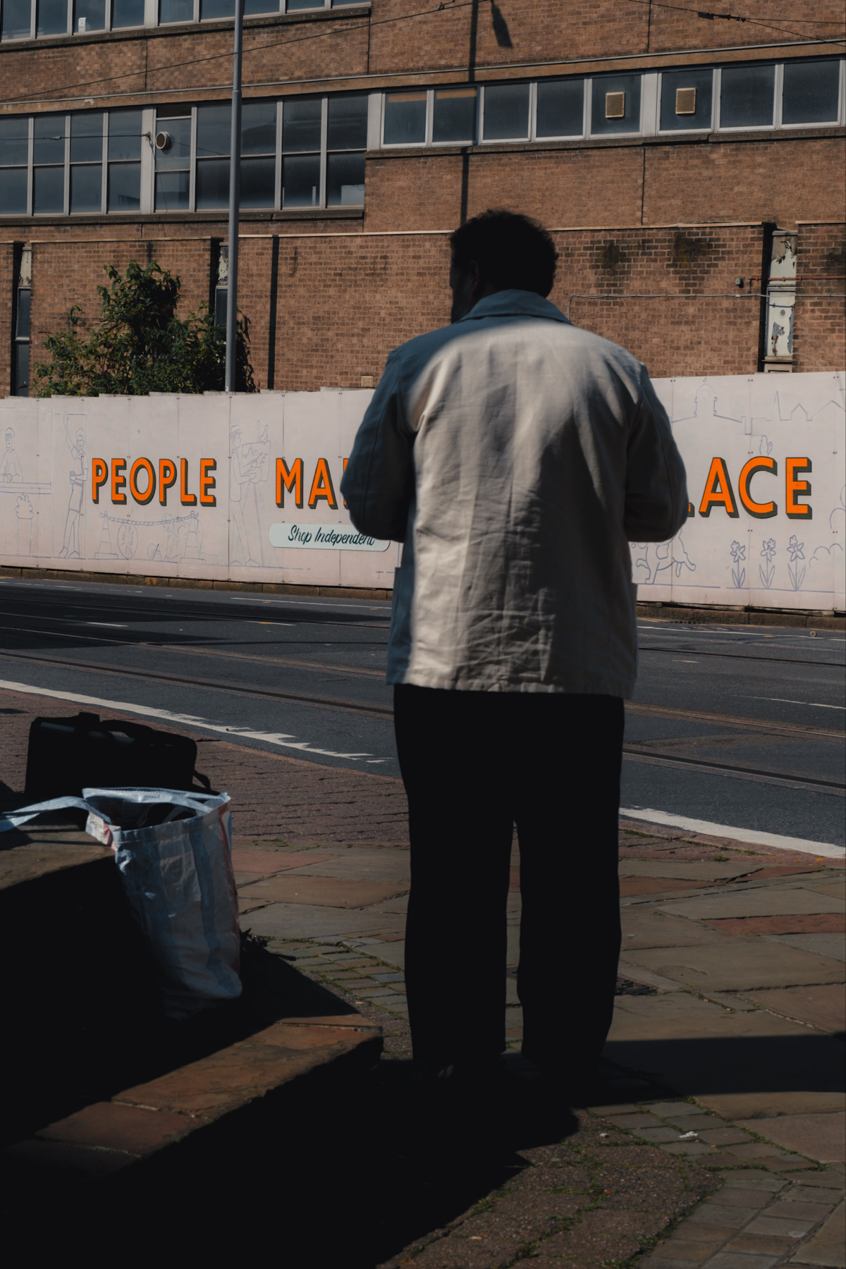 A person in a white jacket stands on a sidewalk, facing away, with their shadow cast on the ground. Bags are placed nearby. In the background, a brick building and a mural with the partial text People A Make Place are visible.