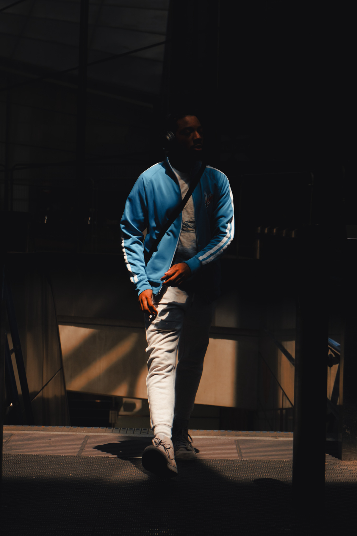 A person walks through a dark, shadowy area, illuminated by a single beam of light. They are wearing a blue jacket, light-colored pants, and sneakers, and they have a crossbody bag. Their face is partially obscured by shadows.