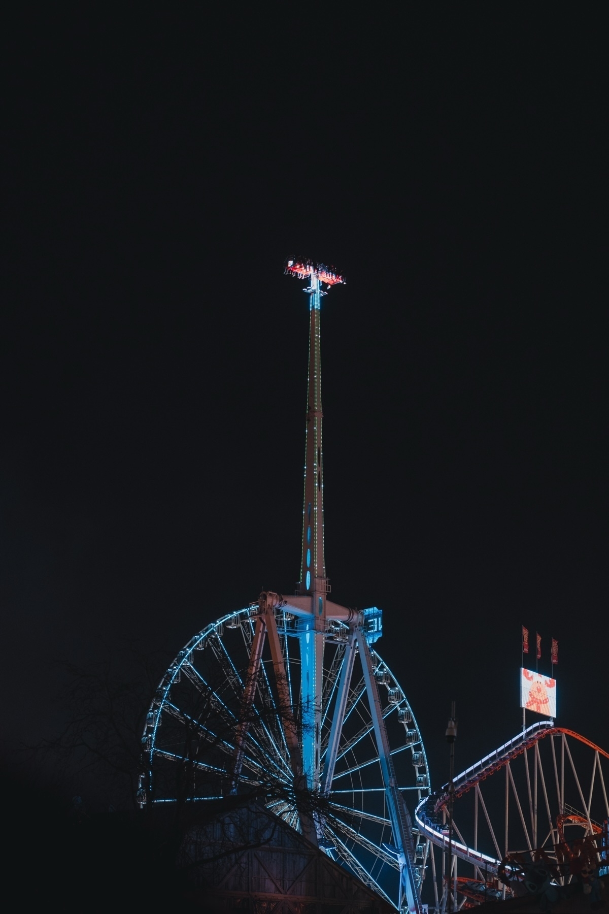 A nighttime amusement park scene featuring a brightly lit Ferris wheel and a tall thrill ride with people on top. The rides are illuminated with blue and red lights against a dark sky, and a roller coaster track is visible nearby.