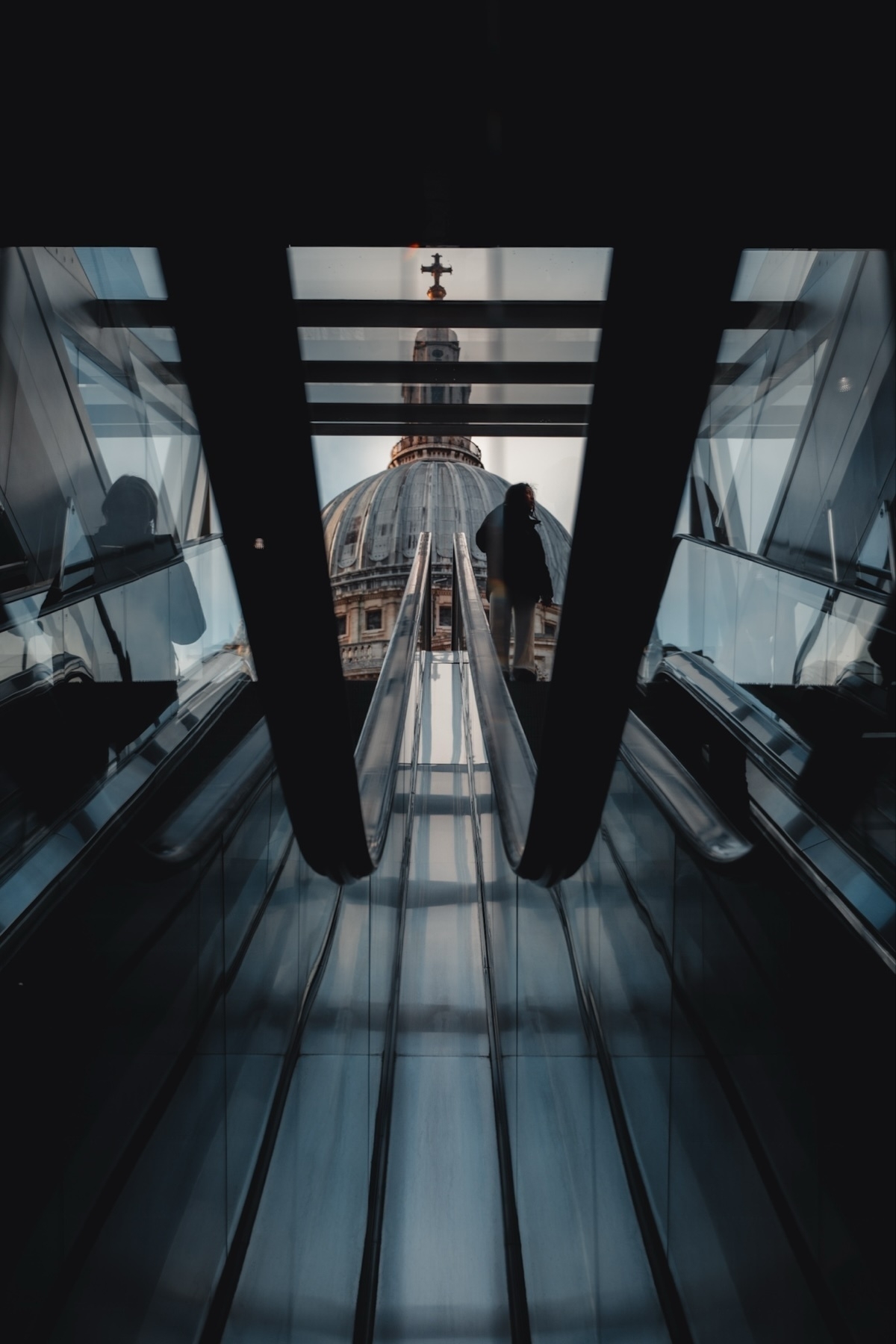 View of an escalator inside a building with a silhouette of a person ascending. In the background, the dome of St. Paul's Cathedral is visible through glass panels. The scene is framed by the dark interior, creating a dramatic contrast with the outside light
