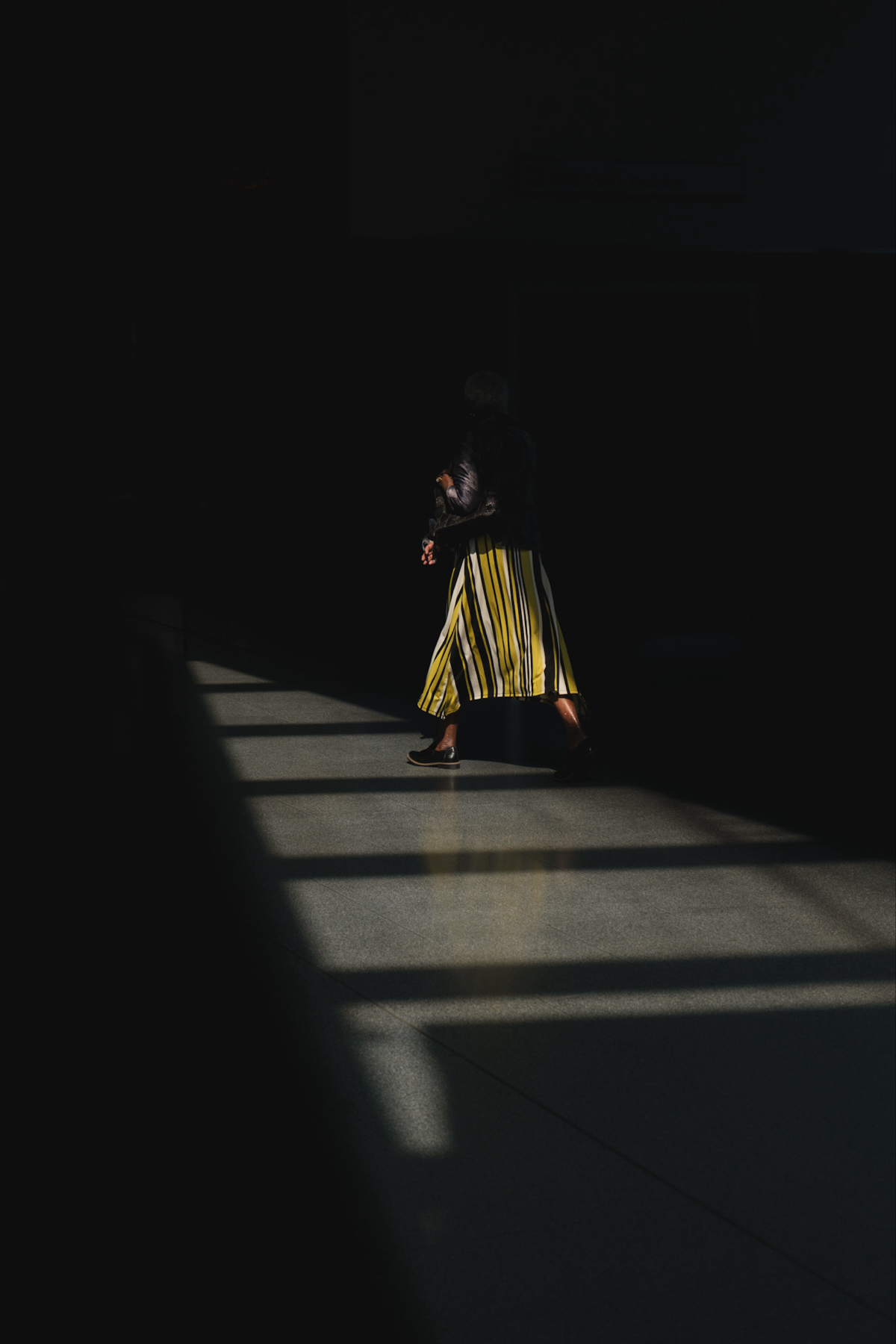 Auto-generated description: A person wearing a yellow and black striped skirt is walking through a dimly lit area with sunlight streaming in through windows.