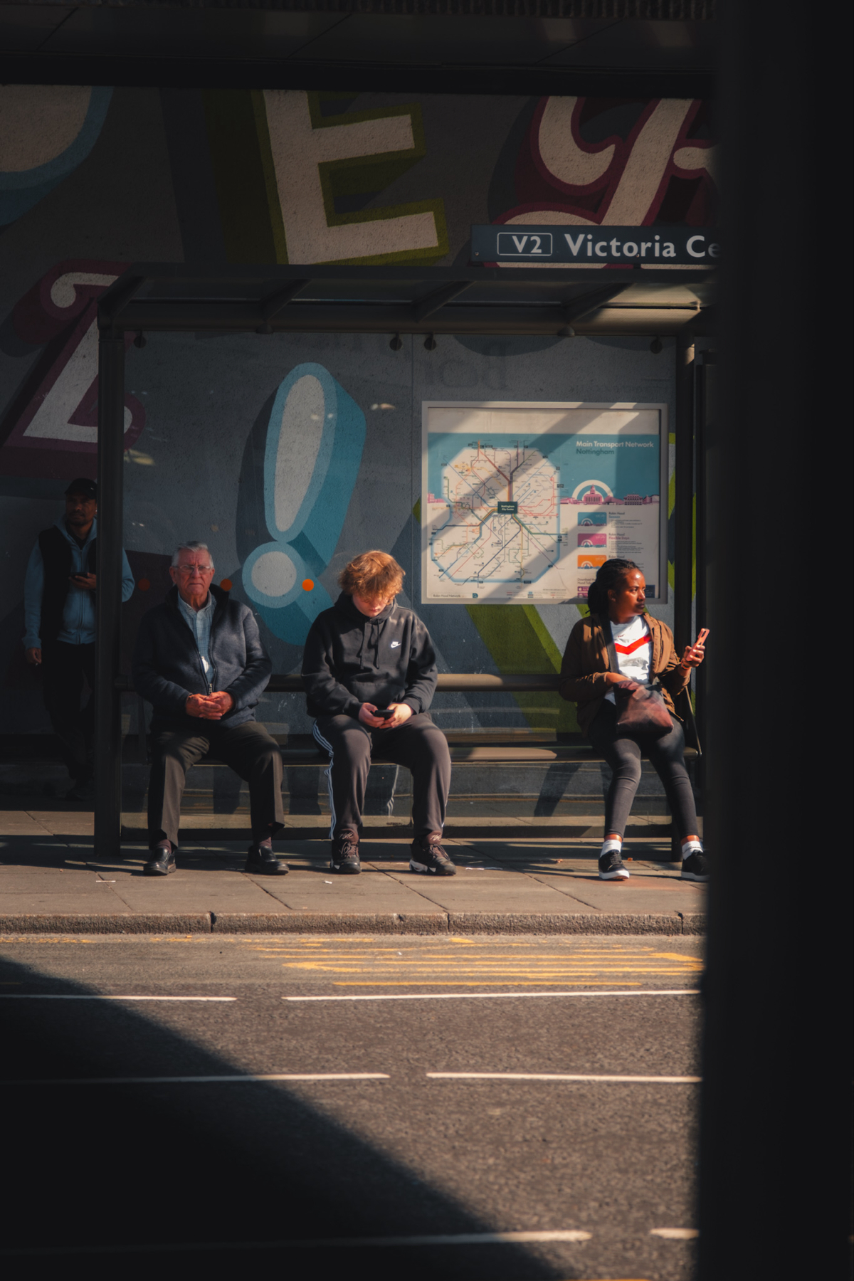 Three people waiting at a bus stop. The bus stop has a sign that reads Victoria Centre and a map of the main transport network. The background features a colorful mural. All three individuals appear to be looking at or holding their phones