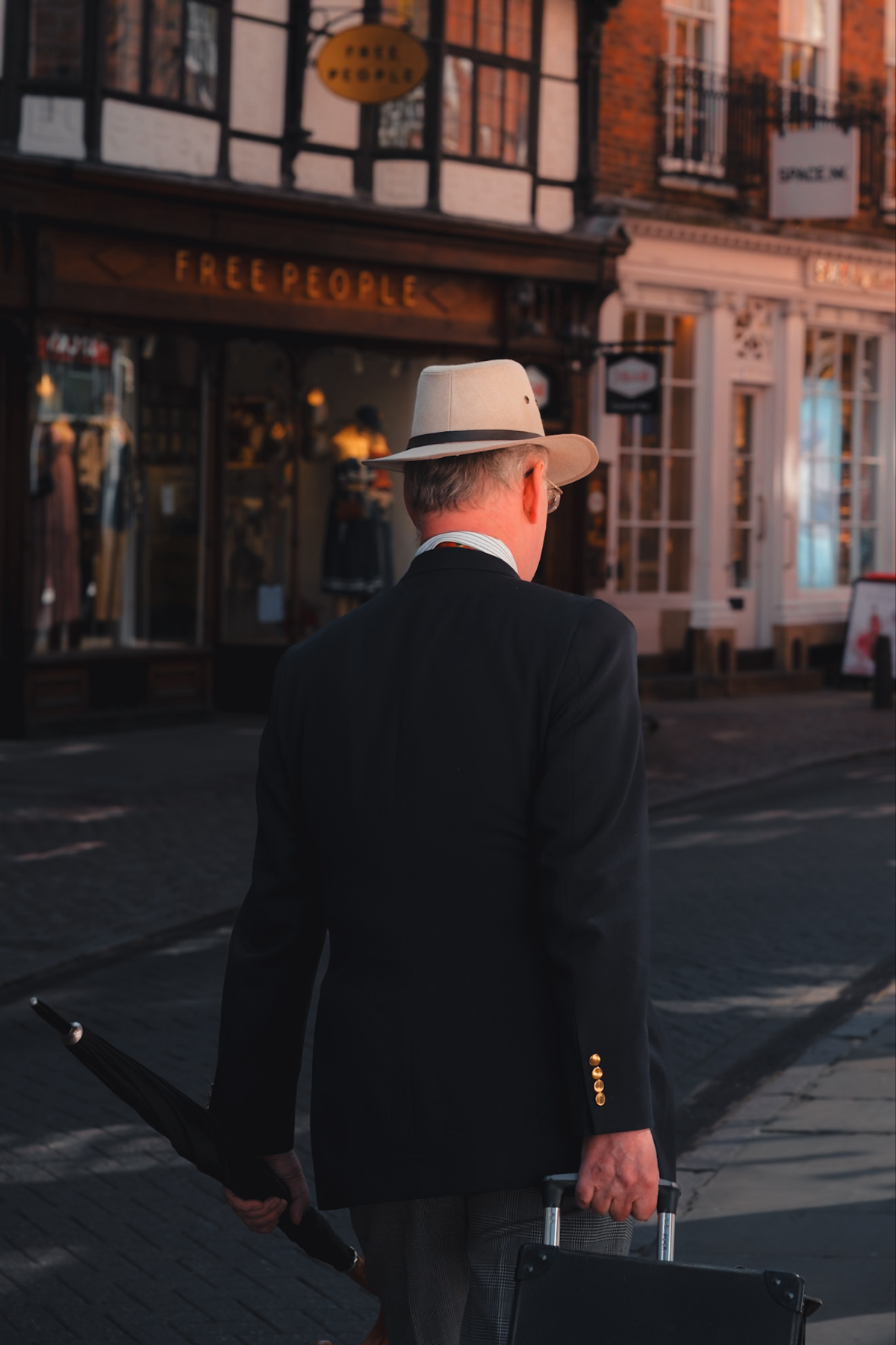 A man wearing a beige hat and dark blazer is walking on the street, carrying an umbrella in one hand and pulling a suitcase with the other. He is seen from the back and is passing by a store with a sign that reads "FREE PEOPLE".