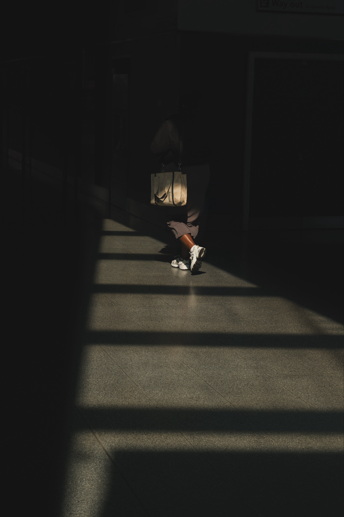 Auto-generated description: A person walks through a dimly lit space with a handbag, illuminated by a slanting beam of light across the floor.