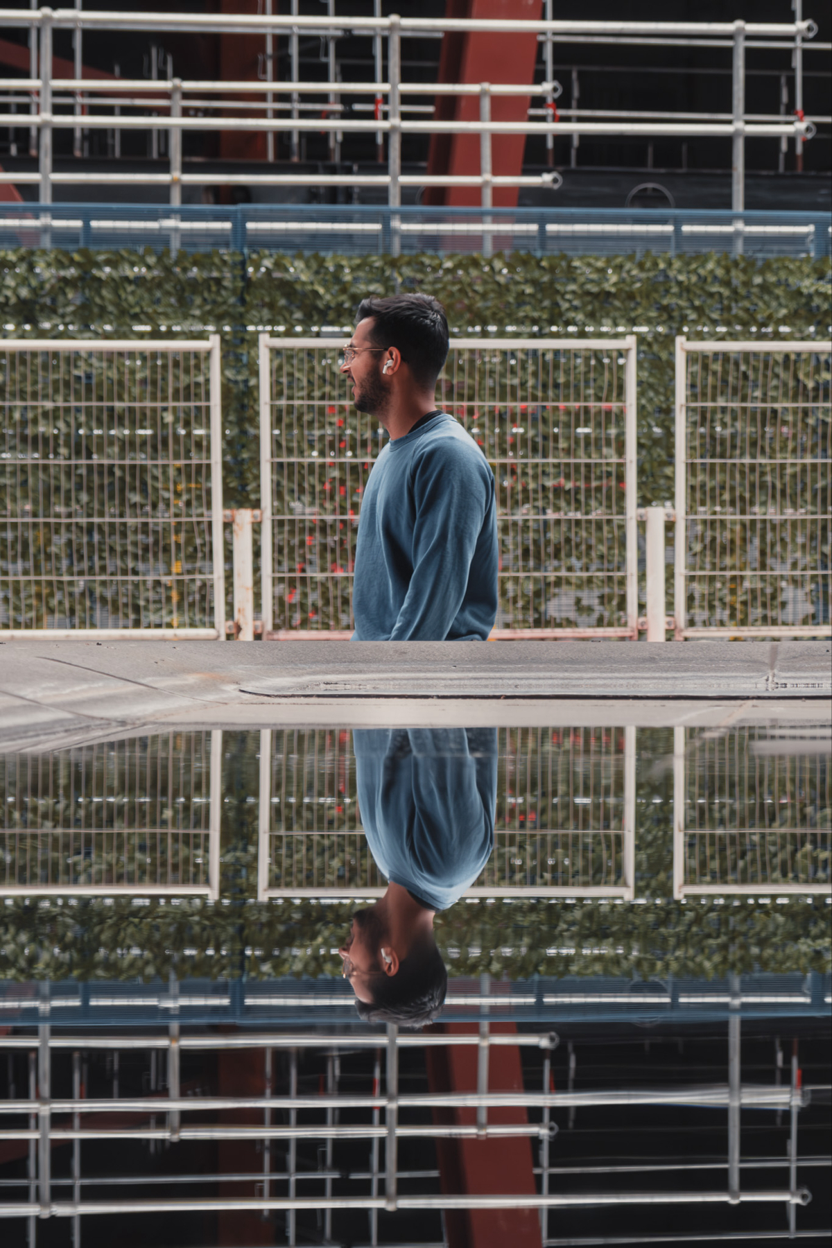 A person wearing glasses and a blue sweatshirt is walking past a construction site or industrial area, reflected in a puddle on the ground.