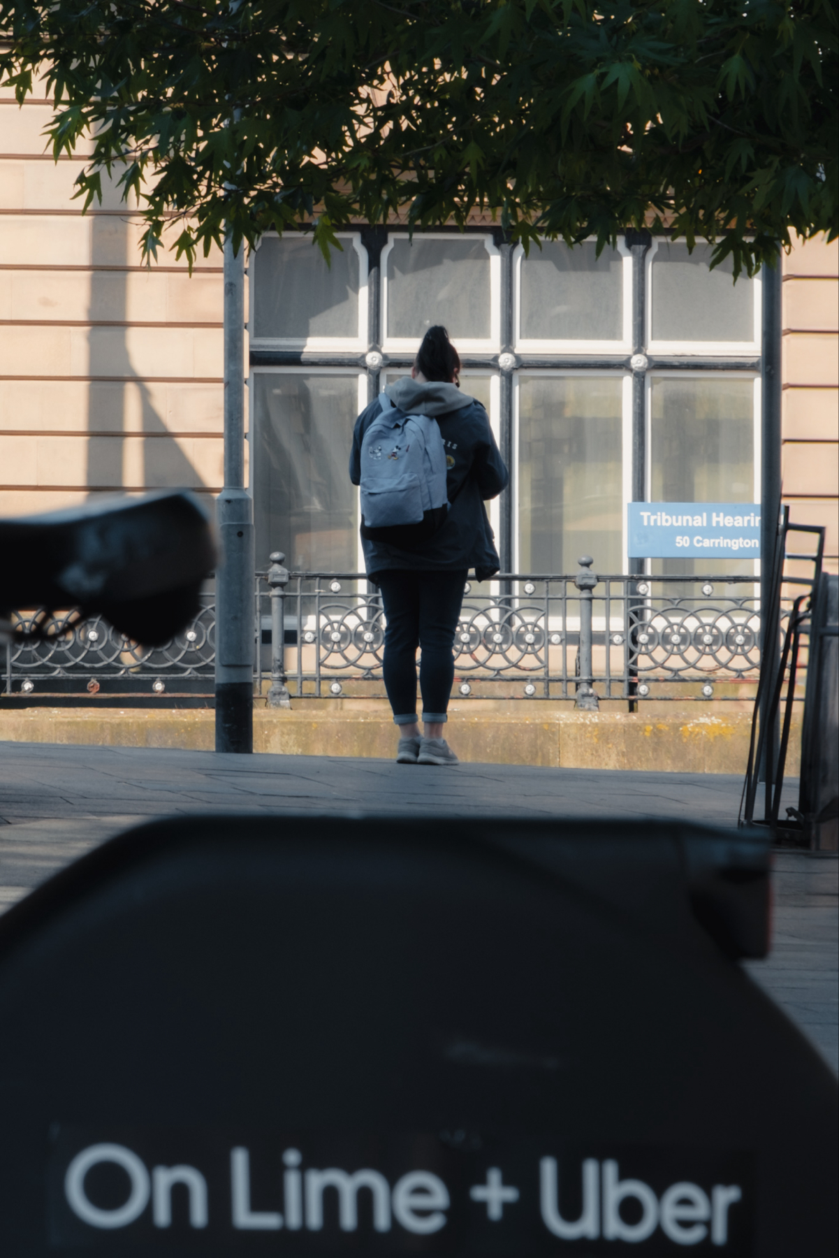 A person wearing a jacket and a light blue backpack is standing on a street in front of a building with a sign that reads Tribunal Hearing 50 Carrington. There is partial text visible in the foreground that reads On Lime + Uber.