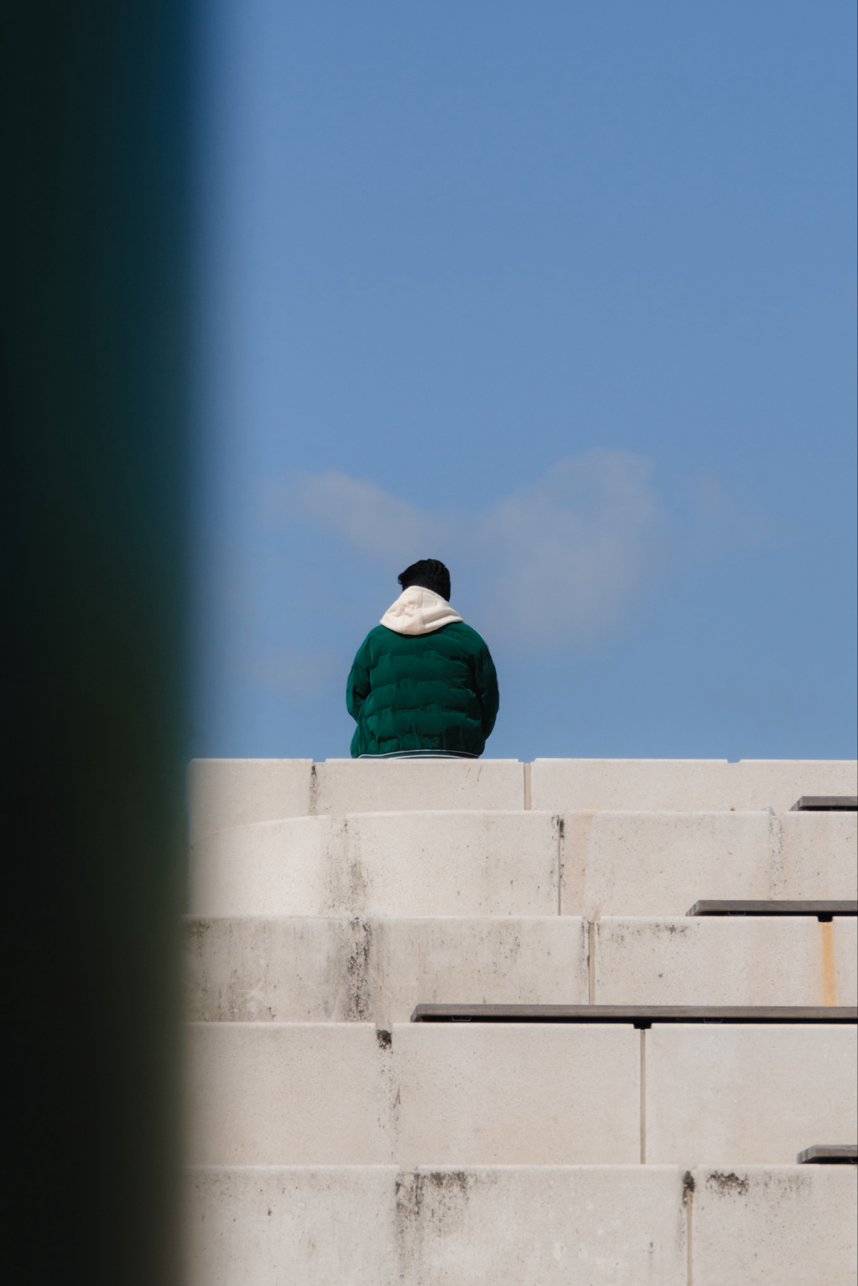 A person in a green jacket with a white hood and a black hat sits alone on concrete steps against a clear blue sky. A vertical green line partially obscures the left side of the image.