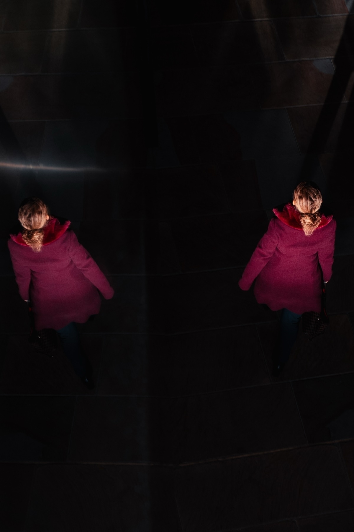A person in a red coat is walking on a dark tiled floor, casting a shadow. The image is taken from above, with dramatic lighting highlighting the coat's color and creating a reflection effect.