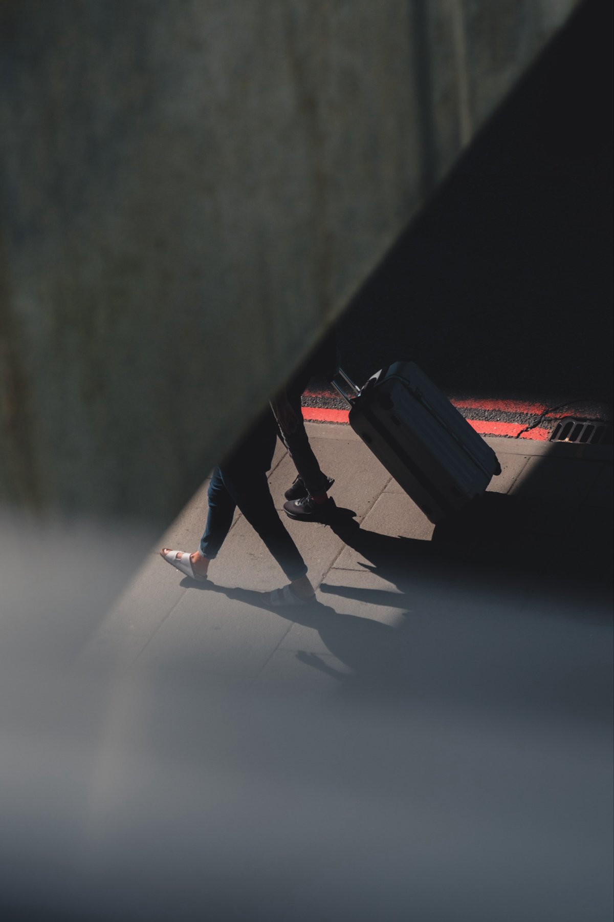 Auto-generated description: A person is walking on a shaded sidewalk, pulling a rolling suitcase.