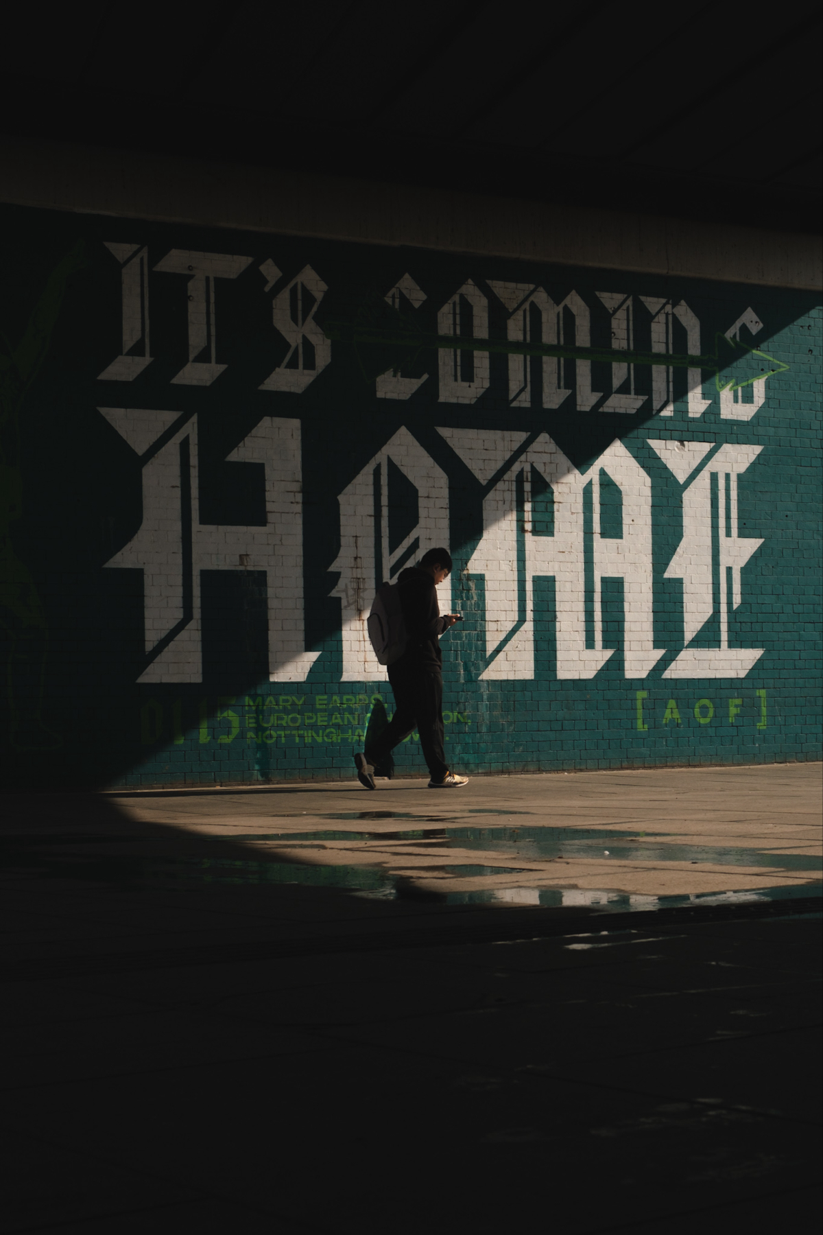 A person is walking under a bridge with graffiti on the wall behind them. The graffiti features large, bold text saying IT'S COMING HOME. The person is wearing a backpack and looking at something in their hand, possibly a phone.