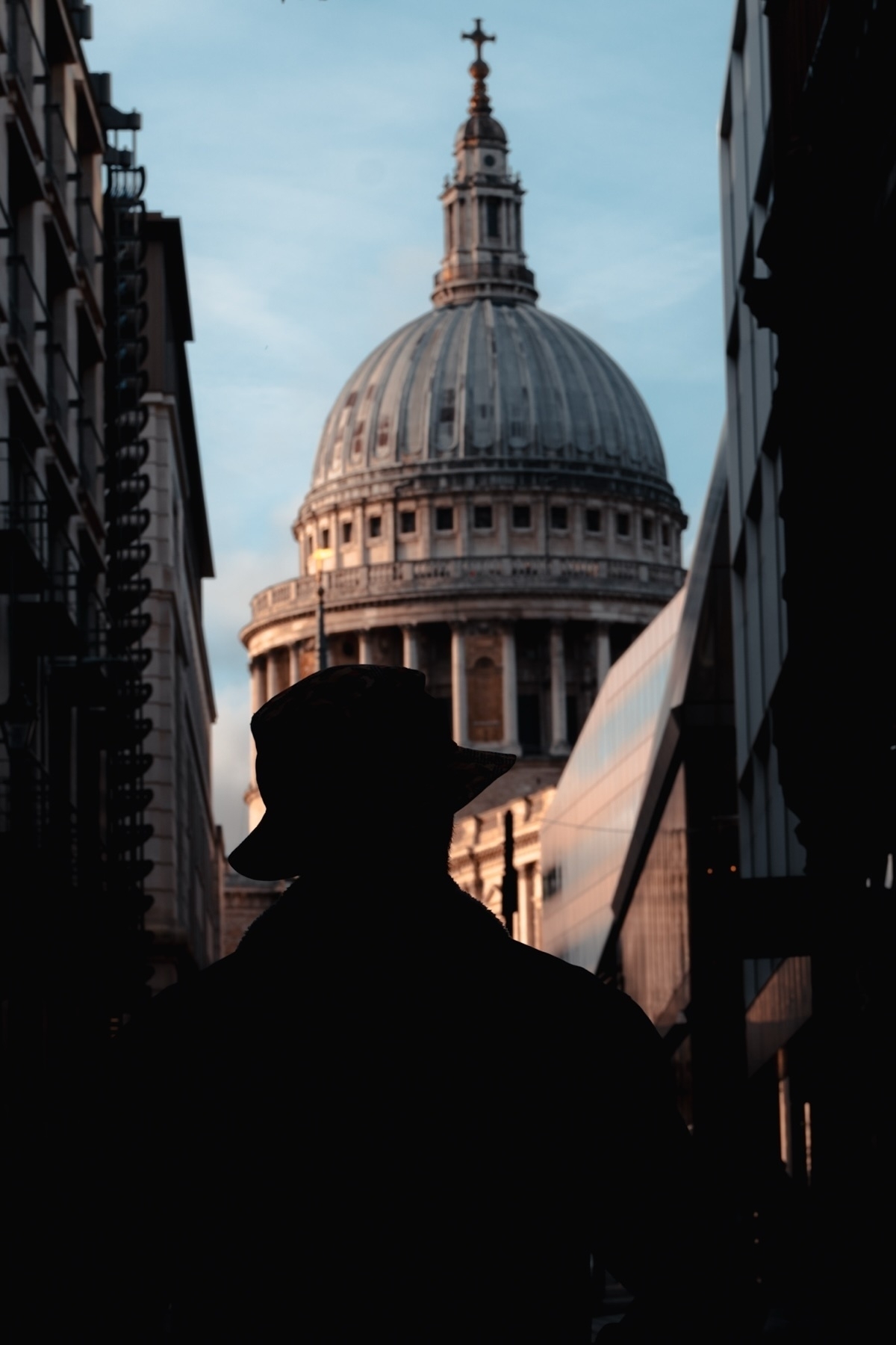 Silhouette of a person wearing a hat, standing in an urban alleyway with St. Paul's Cathedral in the background, under a blue sky.