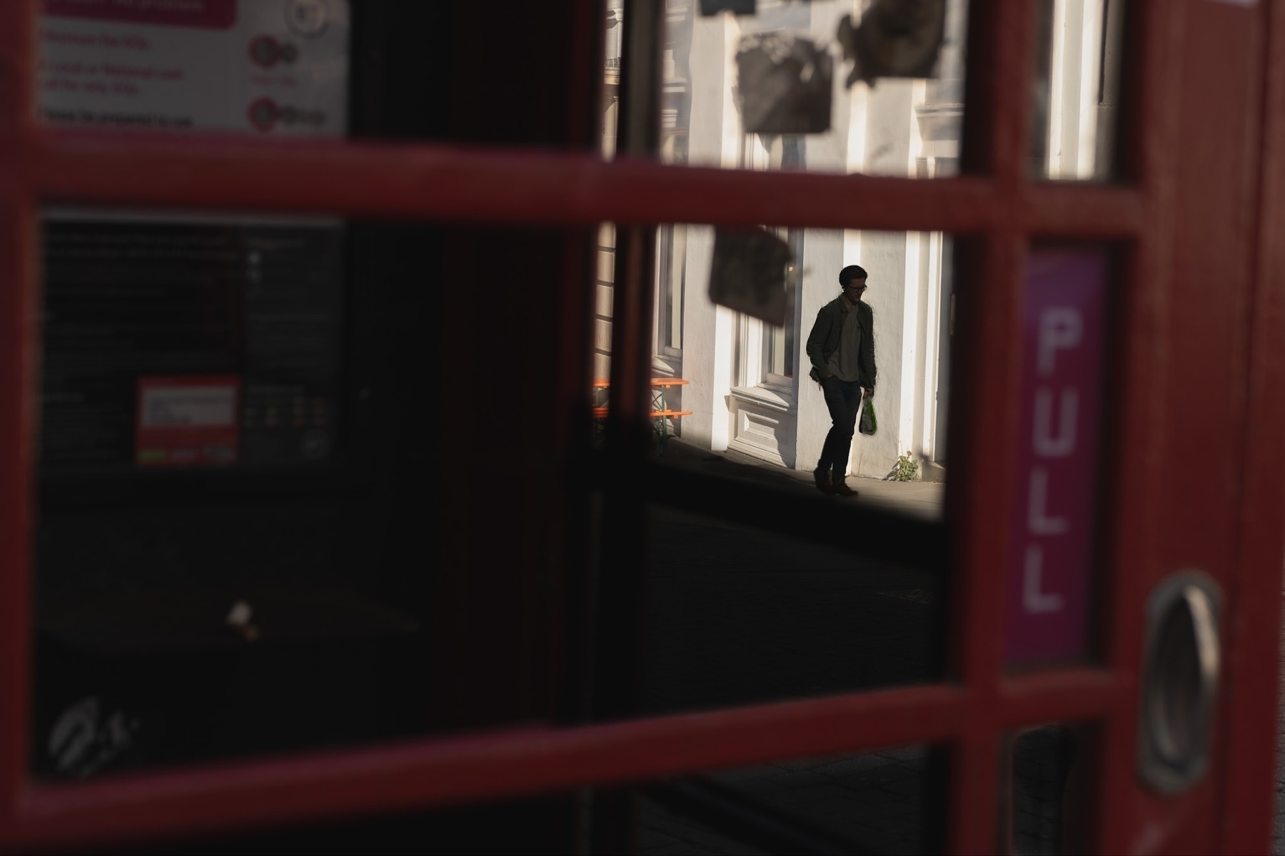 A person is walking outside a building, viewed through the window of a red telephone booth. The word "PULL" is partially visible on the booth's door.