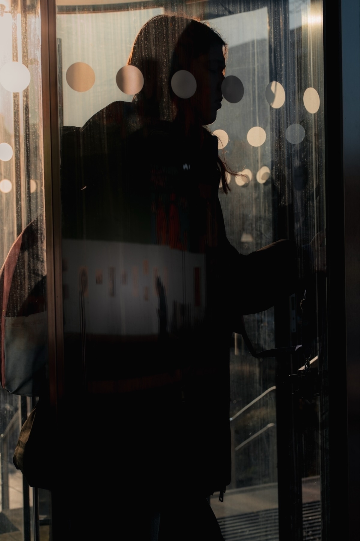 Silhouette of a person entering through a glass door with circular stickers. Sunlight creates reflections and shadows on the glass, adding depth to the scene.