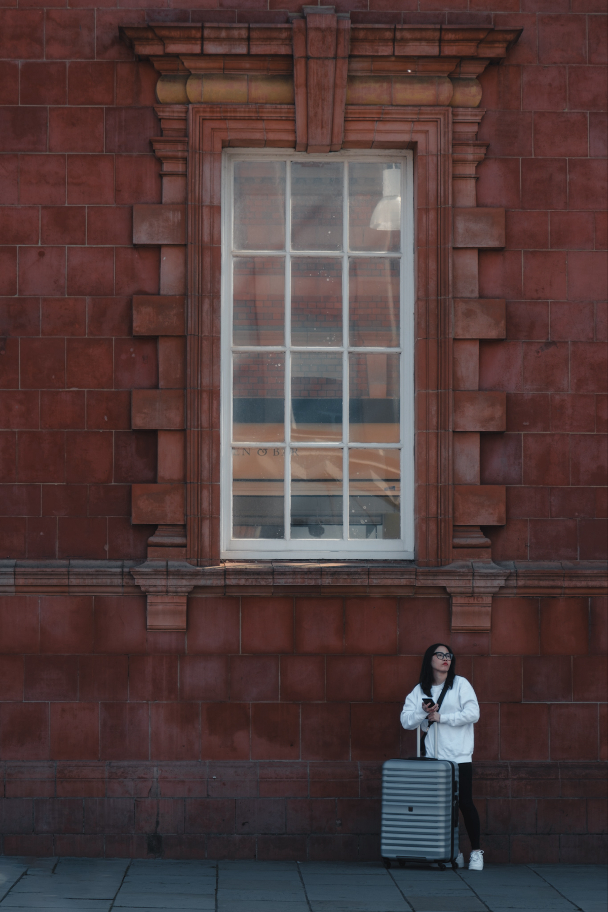 A person stands against a red-brick wall with a large rectangular window. They are holding a smartphone and standing next to a silver suitcase. The person is wearing a white jacket, black pants, and white shoes. The scene appears to be urban.