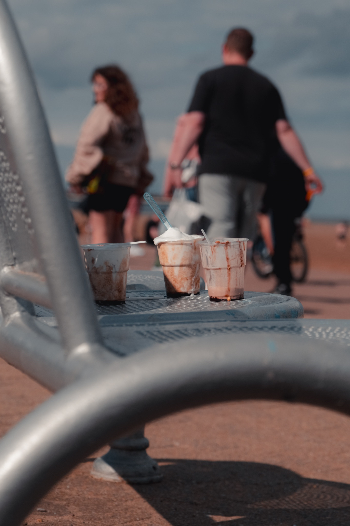 Several partially finished iced coffee cups rest on a metal bench with beachgoers walking by in the background.